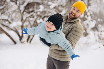 Father holding son like an airplane in a snowy park, both smiling and laughing, enjoying family...
