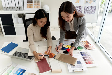 Asian adult women designers collaborate discuss interior design project choosing color material samples architectural plans modern bright office studio professional teamwork creativity planning