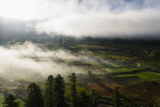 Aerial view of mist-shrouded fields and scattered hamlets, where sunlight pierces through the clouds, painting the valley in hues of gold and emerald, Phobjikha Valley, Wangdue Phodrang, Bhutan.