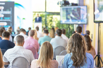 Business conference audience attending keynote presentation in modern seminar hall, corporate event training with speaker on stage and screen