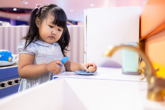 Little Asian girl playing with toy kitchen set in indoor playground,A cute little Asian girl with pigtails focused on playing with a blue toy kitchen set and plastic food in a colorful indoor.