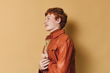 Redhead man wearing brown leather jacket looking sideways with neutral expression on beige...