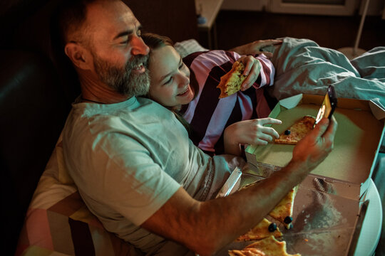 Father and daughter eating pizza in bed while watching smartphone