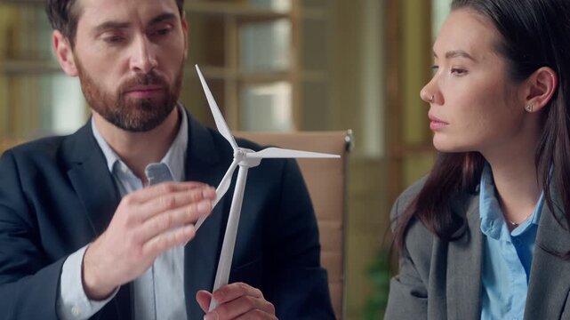 Multiethnic business woman man discussing alternative energy windmill model at office Caucasian businessman Korean businesswoman negotiate explain sustainable source environment ecology collaboration