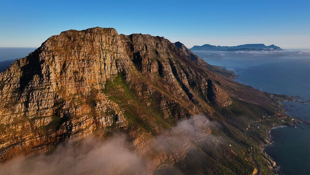 Aerial view of the rugged, sun-kissed cliffs of Table Mountain meet the sapphire Atlantic, a dance of earth and sea veiled in ethereal morning mist, Cape Town, Western Cape, South Africa.