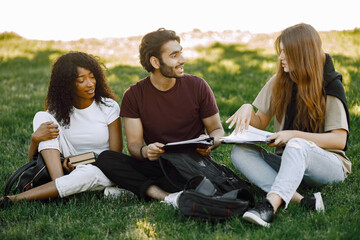 Three international students sitting on a grass in a park and holding a books