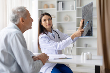 Female doctor holding medical scan and pointing details while consulting senior male patient,...