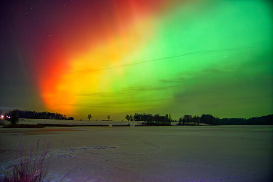 Colorful aurora borealis illuminates a snowy winter landscape under a starry night sky, glowing green, red and yellow over quiet fields.