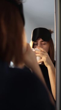 Vertical screen Sleepy woman stands at the mirror with a coffee cup still wearing her sleep mask struggling to focus as she tries to wake up and gather enough energy to start her slow morning