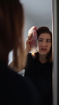 Vertical screen woman holds cold compress near head after minor fall feeling discomfort pain and dizziness while looking at reflection in mirror at home interior