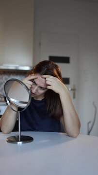 Vertical screen woman presses pimples on face looking into mirror struggling with acne and skin imperfections during stressful morning routine at home feeling frustrated