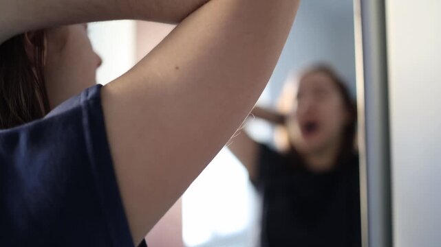 Young woman stands in front of the mirror stretching and yawning still half asleep greeting the early morning light with slow tired movements as she tries to wake up