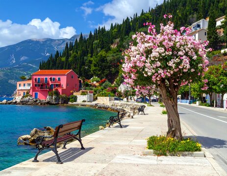 Coastal scene with colorful buildings by the turquoise water. Pink flowering tree and benches line the promenade. Mountains in background