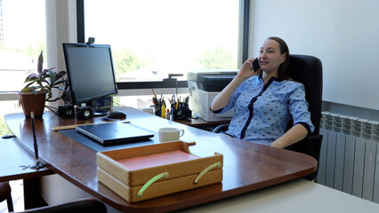 Comfortable office worker chatting on the phone at desk surrounded by computer, stationery, and office equipment during a busy workday