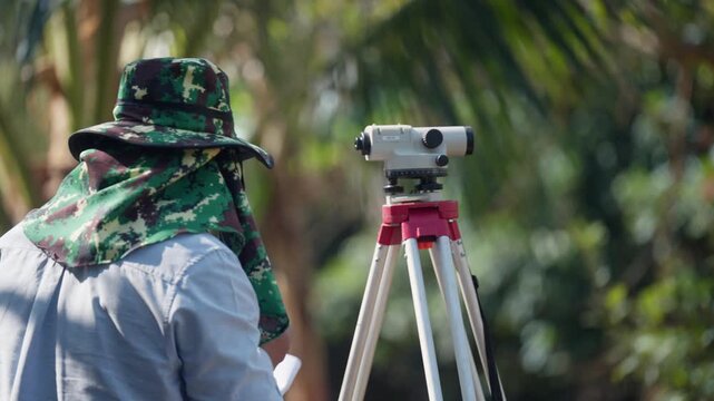 Male civil engineer operating leveling instrument with tripod to conduct a site survey for construction layout planning in tropical outdoor area.