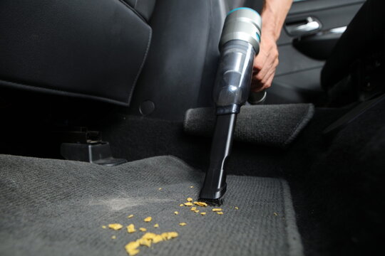 A low-angle close-up shot of a handheld vacuum cleaner using a brush nozzle to suction up yellow food crumbs and hair from a dark grey car floor mat