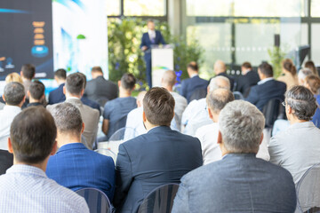 Business audience attending corporate keynote presentation, professional conference seminar with speaker at podium in modern event venue