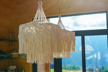 Decorative handmade macrame pendant ceiling lamps hanging in warm wooden interior on large window with mountain view background. Natural light, boho style home decor concept with cozy atmosphere