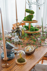 Stylish Easter table decor with candles, greenery, bunny figurines and natural elements in bright spring room interior. Cozy festive dining setup with wooden table, soft daylight and seasonal details