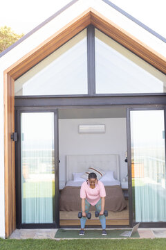 African American woman mid-30s exercising on mat at cabin in pink top lifting weights with laptop