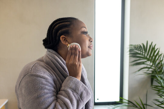 Mid adult African American woman applying toner with cotton pad at bathroom LED mirror wearing robe