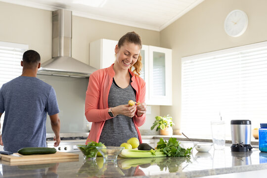 Couple cooking and prepping in kitchen, woman peeling fruit at stone counter wearing coral jacket