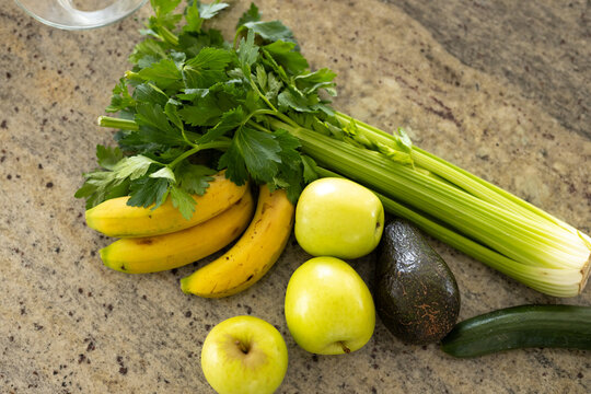 Celery, bananas, green apples, avocado and cucumber resting on granite counter, glass rim visible