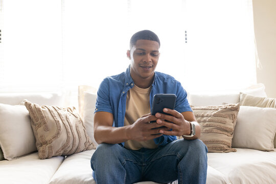 African American man sitting on light sofa at home wearing blue shirt holding dark smartphone