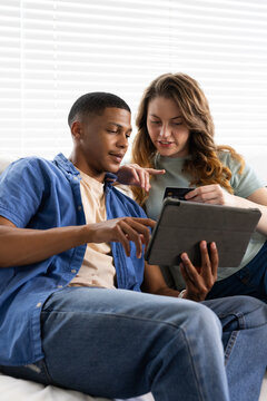 Diverse couple sitting on sofa at home, wearing casual clothes, holding tablet and card, pointing