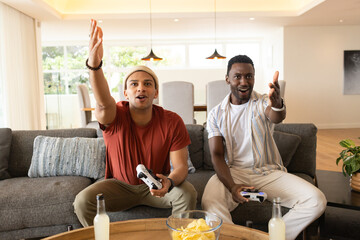 Diverse male friends sitting on gray sofa, holding controllers, sharing chips on coffee table