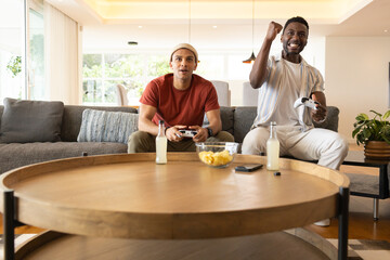 Male friends in red tee, striped shirt sitting in living room on sofa gaming with controllers