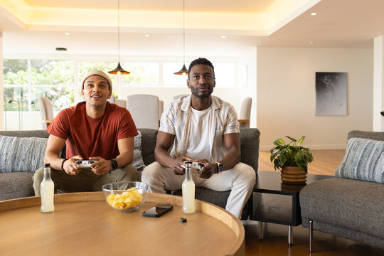 Diverse male friends sitting on gray sofa in living room playing games with controllers and chips