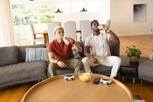 Diverse male friends sitting on gray sectional sofa in living room drinking from glass bottles