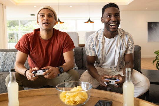 Diverse male friends playing console game on sofa in living room with controllers chips drinks