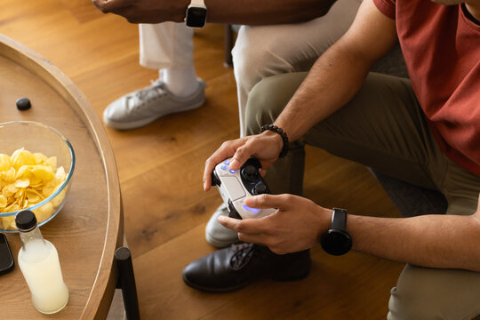 African American man with male friend sitting in lounge holding game controller near chips on table
