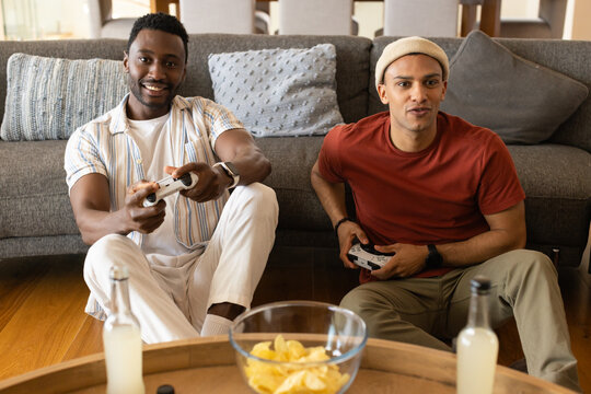 Diverse male friends sitting on wood floor in living room playing with controllers, sharing chips