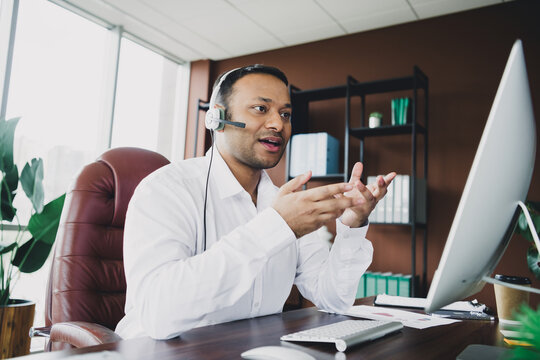 Businessman in an office headset explains plans during video call at modern loft office setup large computer screen and busy desk professional