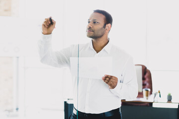 Young handsome businessman presents in a modern office using a marker on a glass board while holding a document and thinking about strategy