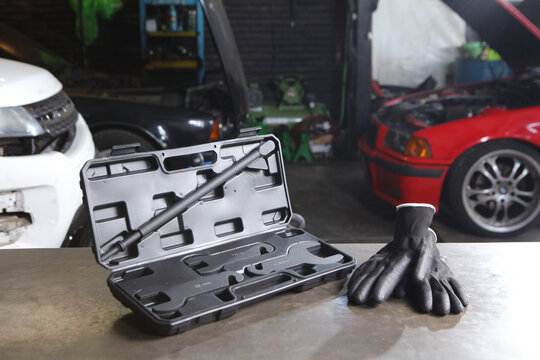 An open black plastic tool case containing specialized automotive wrenches and a long metal rod sits on a workbench next to a pair of black work gloves in a garage with cars.