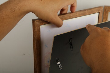 Hands are shown placing a black backboard into a wooden picture frame over a piece of artwork, demonstrating the assembly process of a professional-style home decor wall frame.