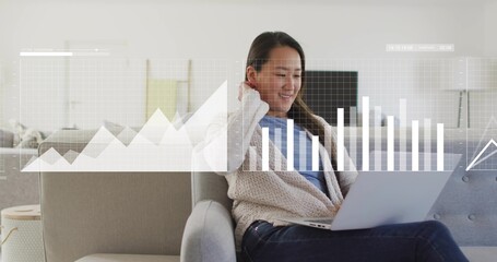 Analyzing woman wearing beige cardigan and dark jeans, typing on laptop in living room with charts