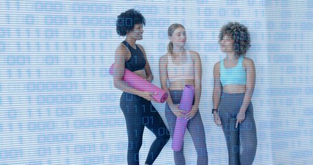 Standing trio in sportswear holding pink and purple rolled yoga mats in yoga studio, fitness watch