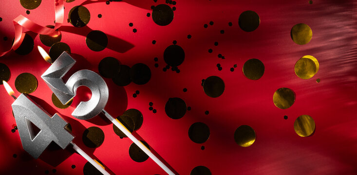 Concept of celebrating birthday and anniversary. Number 45 forty-five silver celebration birthday candle on festive red background with large golden confetti. Forty-five years birthday.