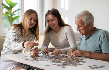 Fototapeta premium Sisters And Grandpa Having Fun, Connecting A Puzzle And Laughing Together