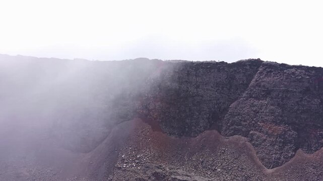 Drone footage at the edge of a volcanic crater at Piton de la Fournaise on Reunion Island. Steep crater walls and mineral textures. Wave of fog drifts in from the left. Hikers are barely visible.