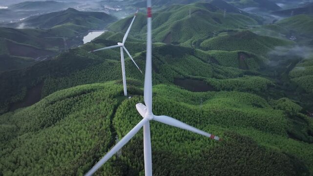 White wind turbines standing on green hills with dense forest and natural landscape captured from aerial view
