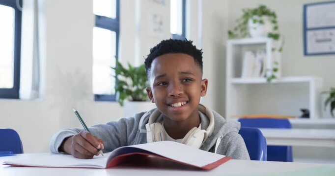 African teenage male at desk writing with pencil in red notebook studying gray hoodie headphones