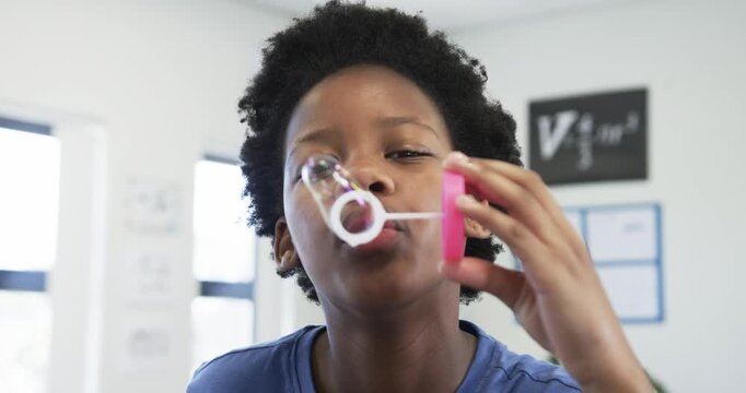 African American child dipping ring wand in pink tub and blowing stream, making bubble in classroom