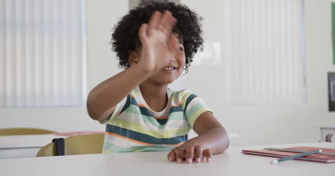 African American child reacting to off-camera voice waving smiling fidgeting at desk in classroom