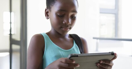 African American child standing in bright hall with bag holding tablet, tapping to do homework - Powered by Adobe